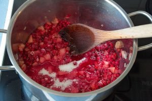 Wooden spoon stirring lingonberries and apples in saucepan for jam.