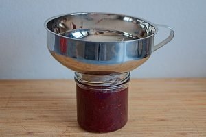 Hot lingonberry jam being poured into a sterilised jar through a funnel.