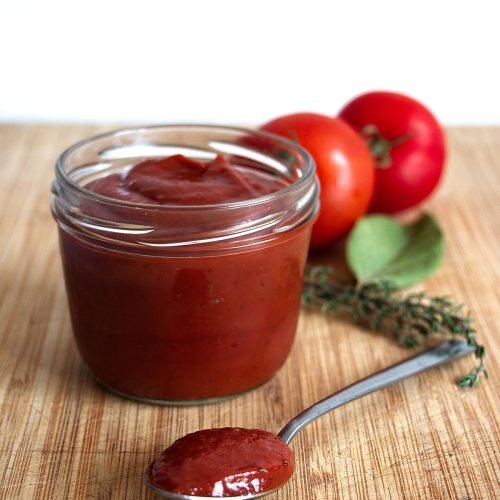 Homemade tomato ketchup in a glass jar with a spoonful on wooden board, fresh tomatoes and thyme in background