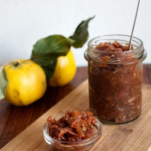 Jar and bowl of homemade Caramelised Onion and Quince Chutney on a wooden board with fresh quinces.