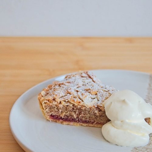 Slice of lingonberry Bakewell tart on a plate, served with whipped cream.