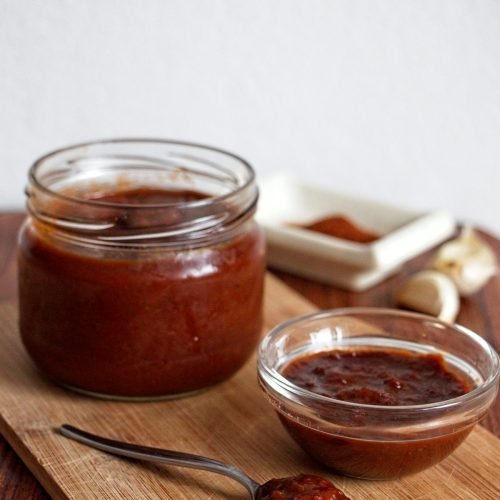 Homemade Cornelian Cherry BBQ Sauce in a jar and bowl with a spoon on a wooden board.