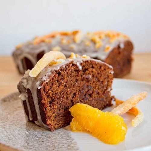 Slice of gingerbread with orange glaze served with candied peel and fresh orange, loaf in the background