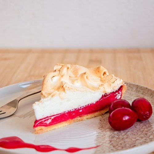 Slice of cornelian cherry meringue pie on a white stoneware plate with cherries and syrup