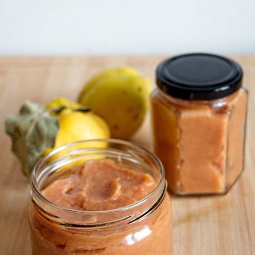 Two jars of homemade quince butter on a wooden board with fresh quinces in the background.