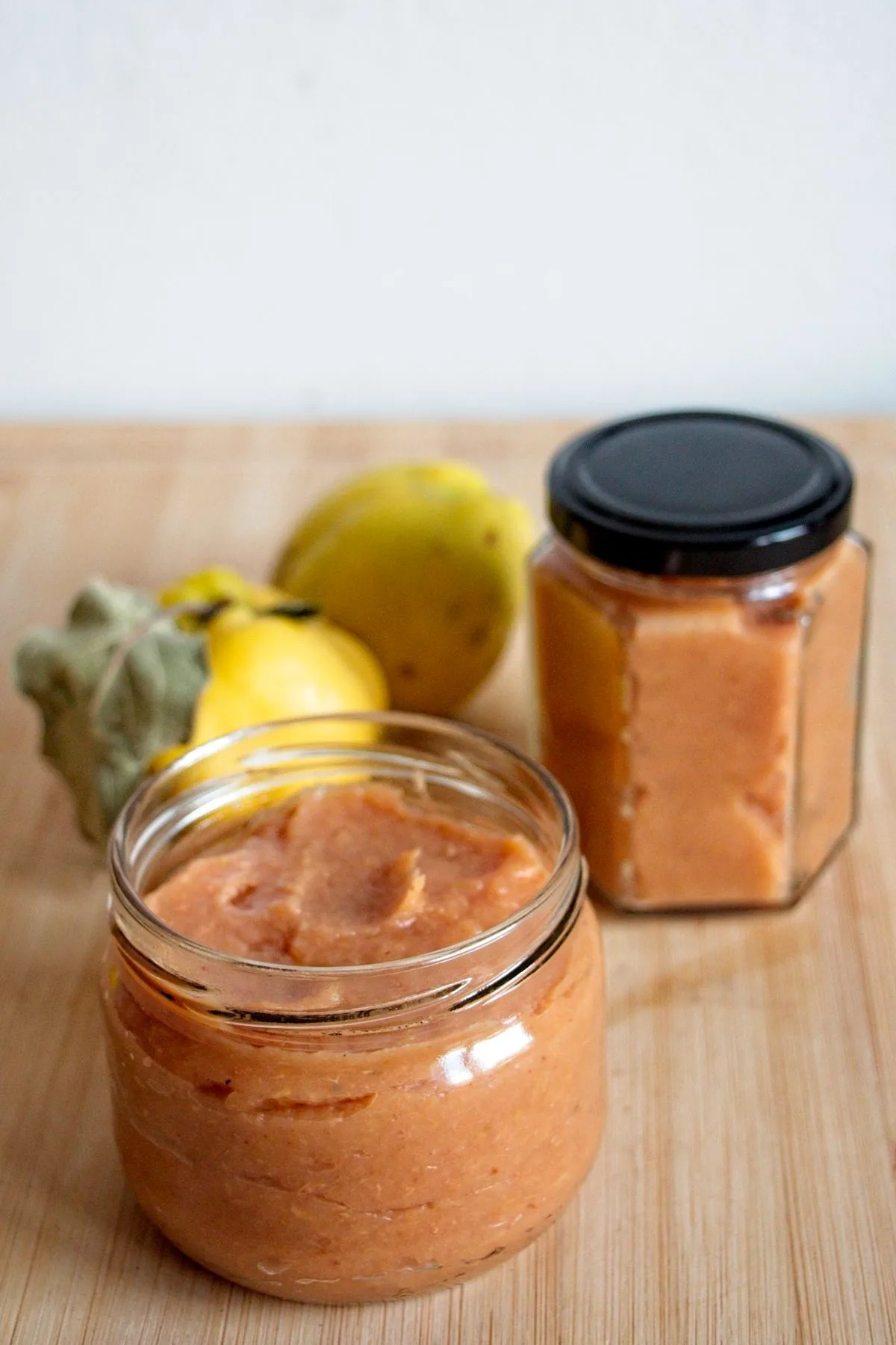 Two jars of homemade quince butter on a wooden board with fresh quinces in the background.