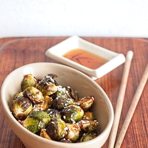 Bowl of miso-roasted Brussels sprouts with sesame seeds, served beside a small dipping dish and chopsticks.