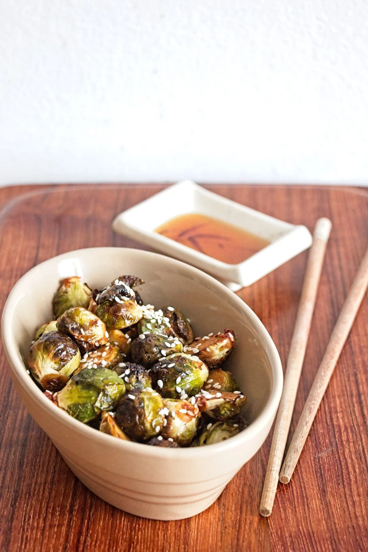 Bowl of miso-roasted Brussels sprouts with sesame seeds, served beside a small dipping dish and chopsticks.