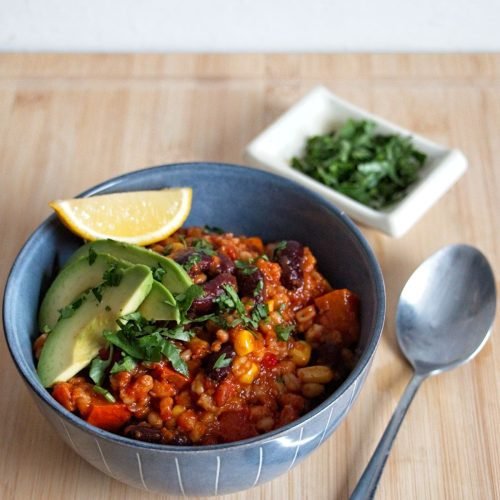 Bowl of Pumpkin Wheat Berry Chili topped with avocado and a lemon wedge, with herbs and a spoon on a wooden board.