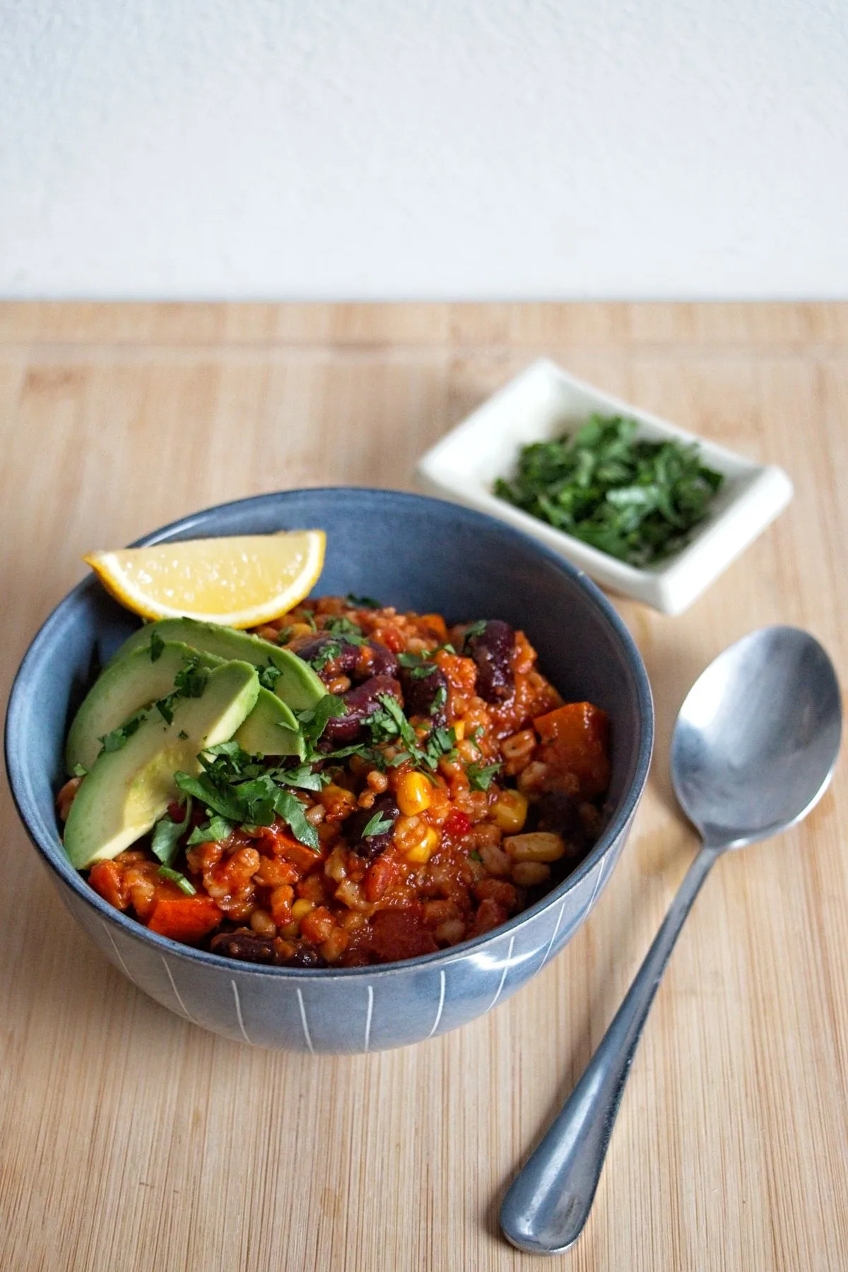 Bowl of Pumpkin Wheat Berry Chili topped with avocado and a lemon wedge, with herbs and a spoon on a wooden board.