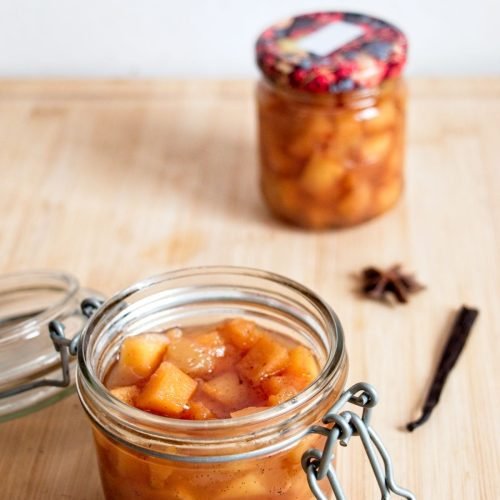 Jar of quince compote with vanilla and cardamom, surrounded by spices on a wooden surface.