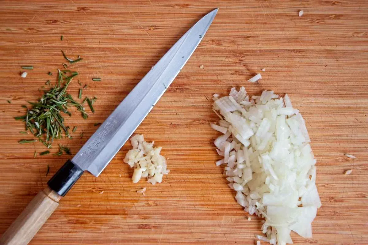 Chopped onion, garlic, and rosemary on a wooden board with a knife.