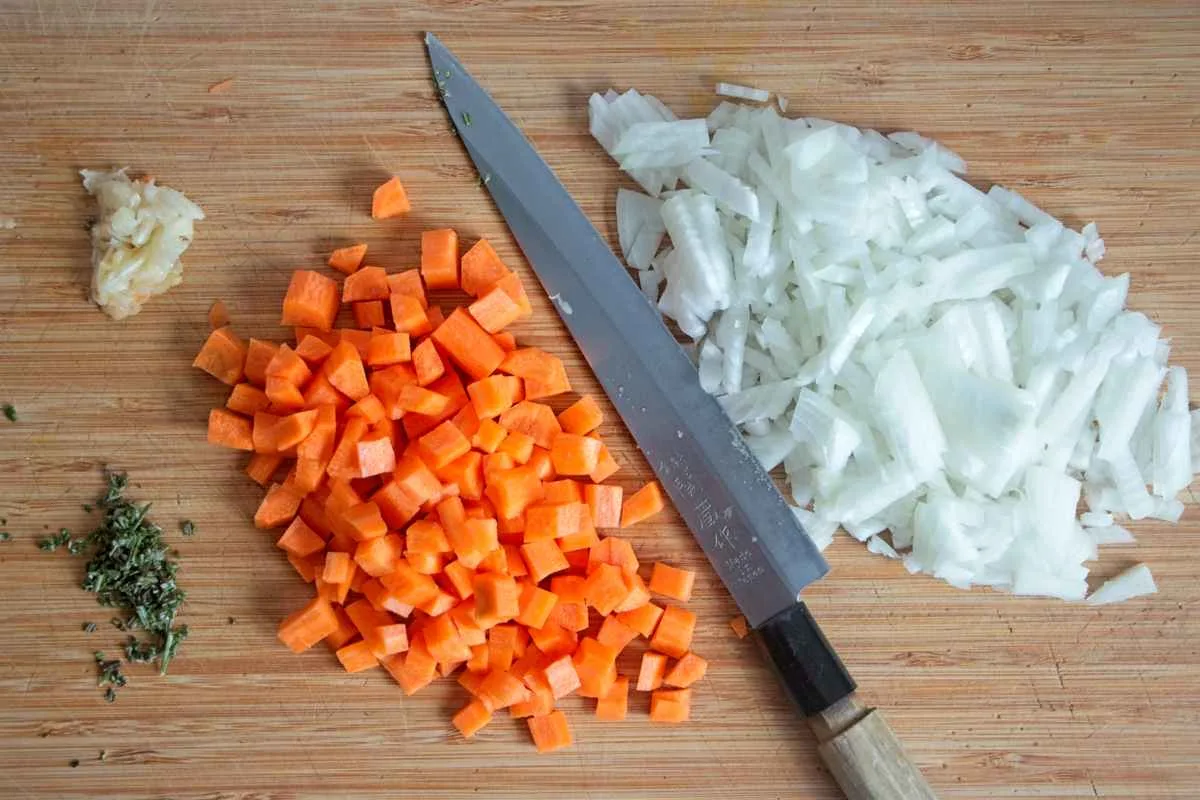 Chopped carrots, onions, garlic, and herbs on a wooden board with a knife ready for vegetarian cottage pie with porcini