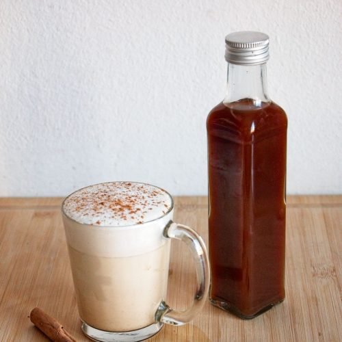 Homemade spiced chai syrup in a glass bottle beside a chai latte topped with foam and cinnamon.