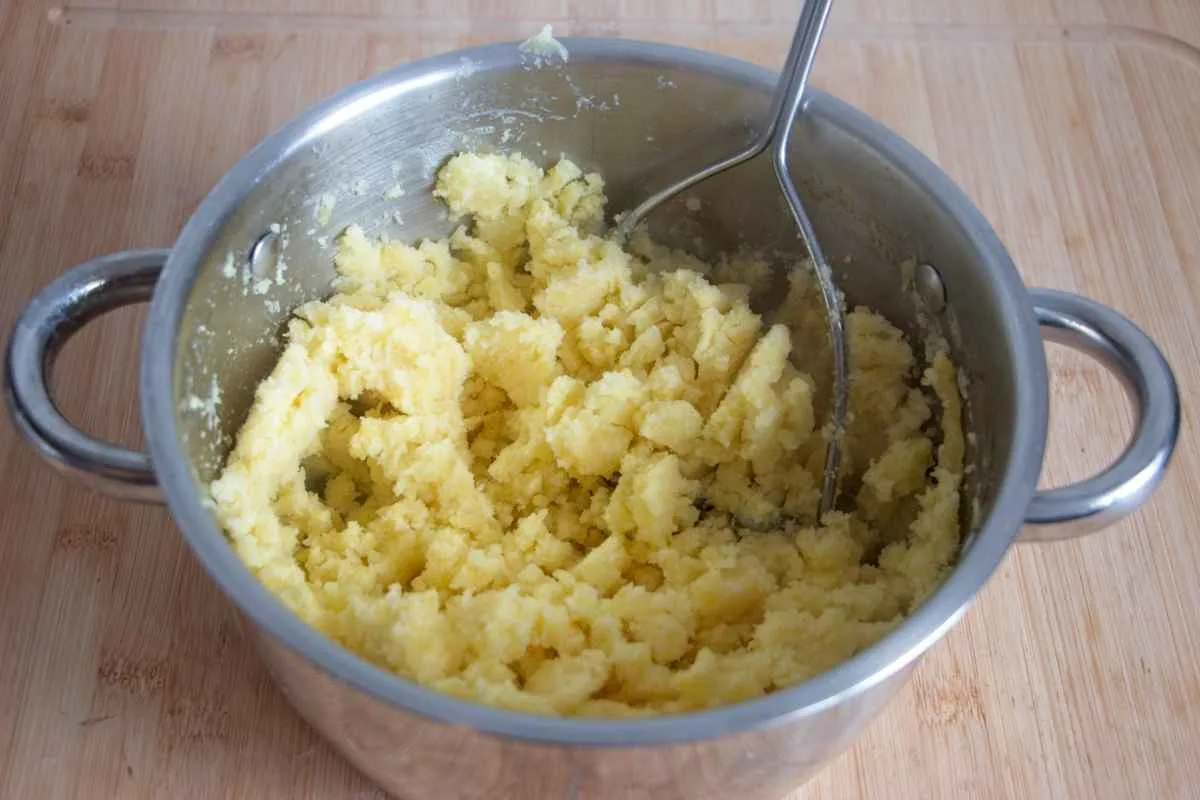Partially mashed potatoes in a saucepan with a masher.