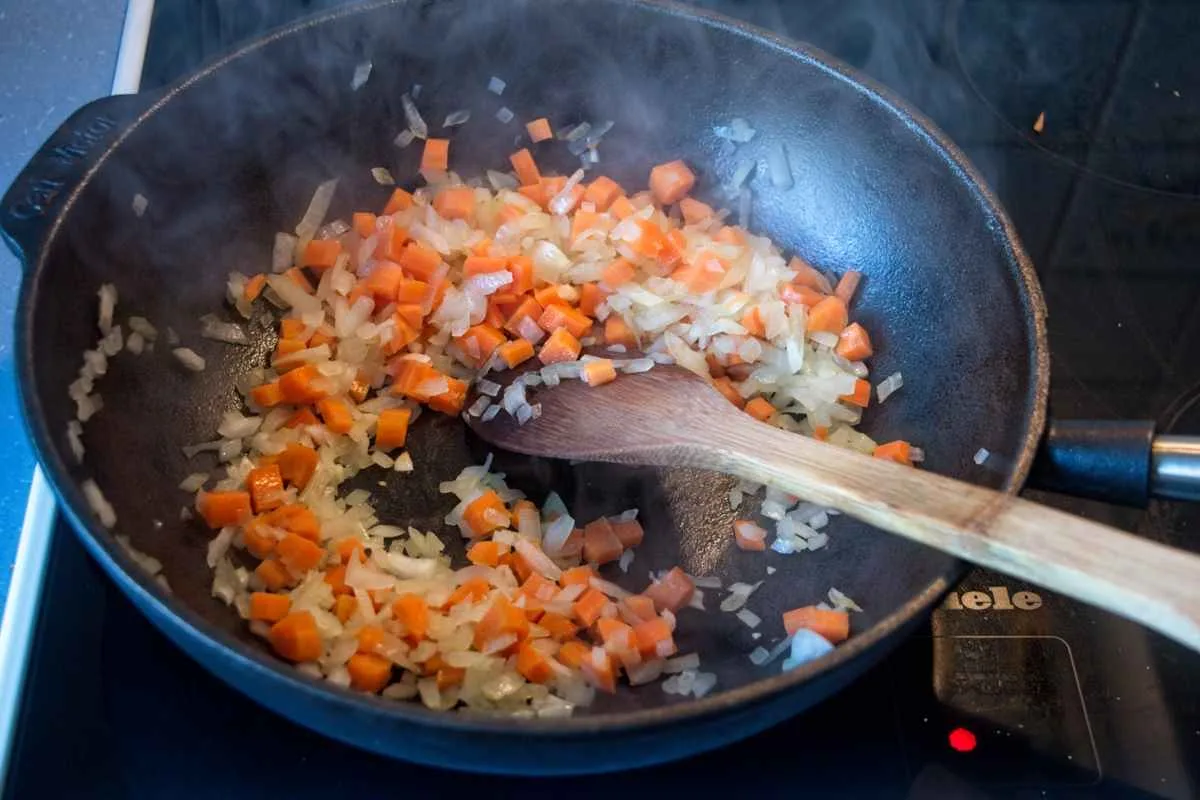 Onions and carrots sautéing in a pan with a wooden spoon, steam rising.