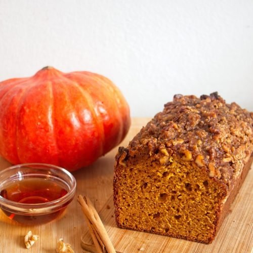 A pumpkin spice and maple loaf with a walnut crumble topping sliced open on a wooden board, with a pumpkin, maple syrup, walnuts, and cinnamon beside it.