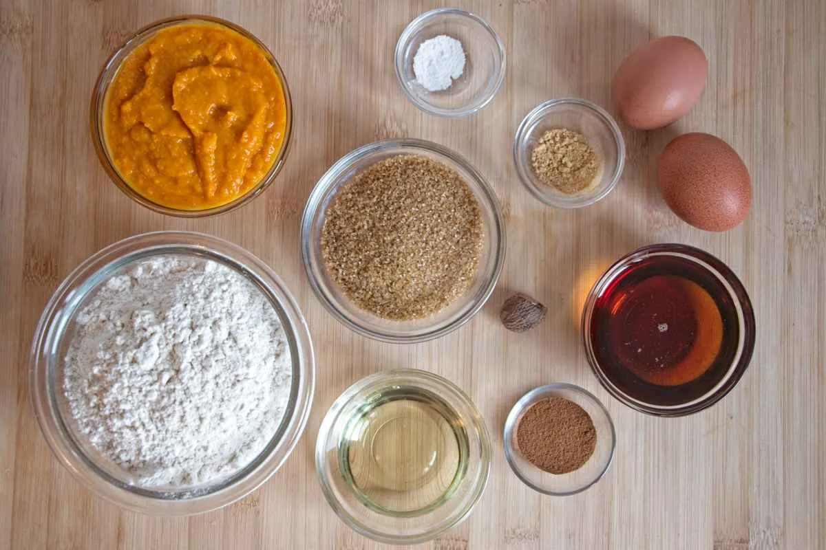 Ingredients for a pumpkin spice and maple loaf arranged in bowls, including pumpkin purée, flour, sugar, eggs, spices, and oil.