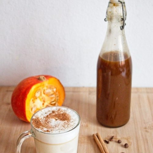 Homemade pumpkin spice syrup in a glass bottle beside a mug of spiced latte with cinnamon and nutmeg.