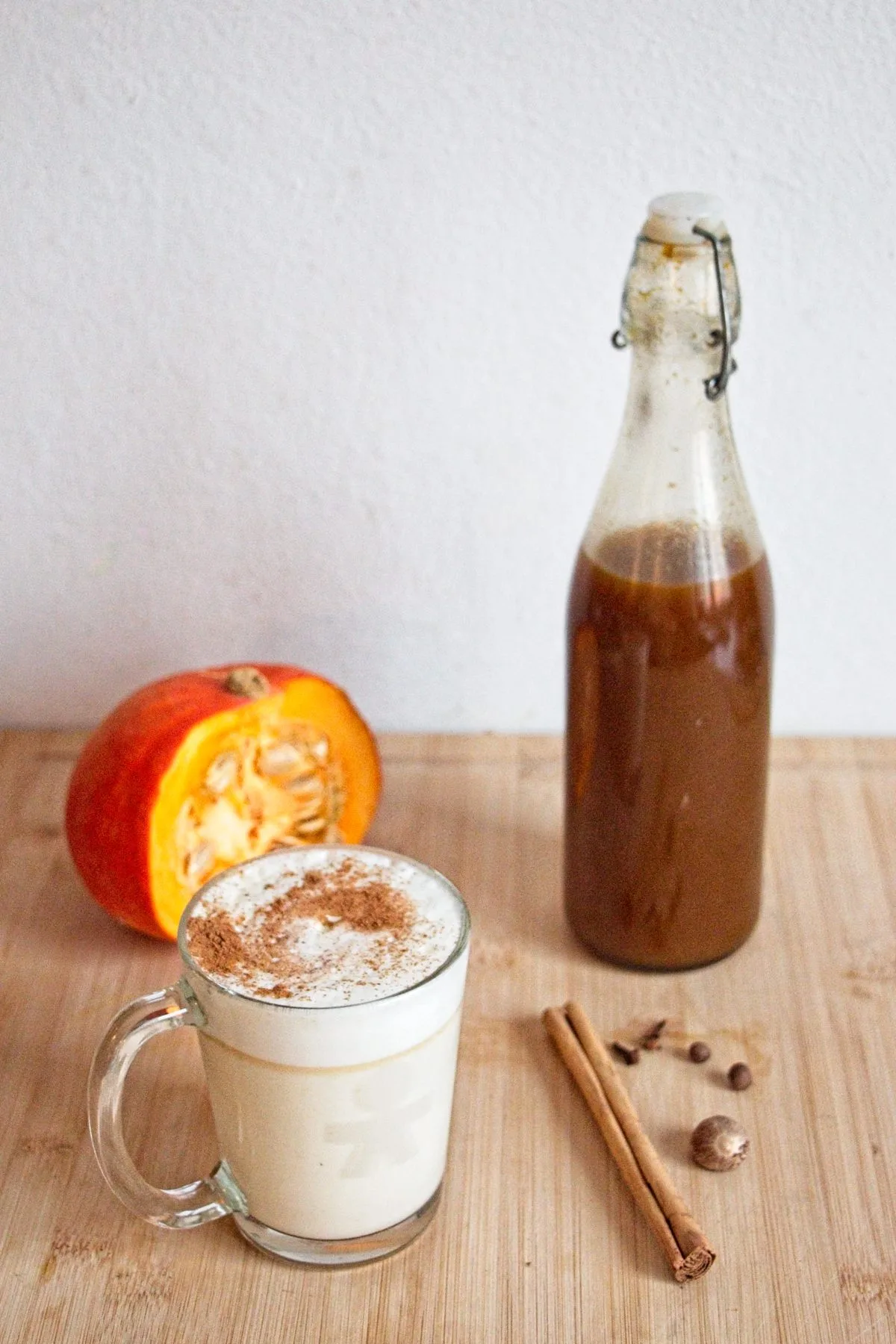 Homemade pumpkin spice syrup in a glass bottle beside a mug of spiced latte with cinnamon and nutmeg.