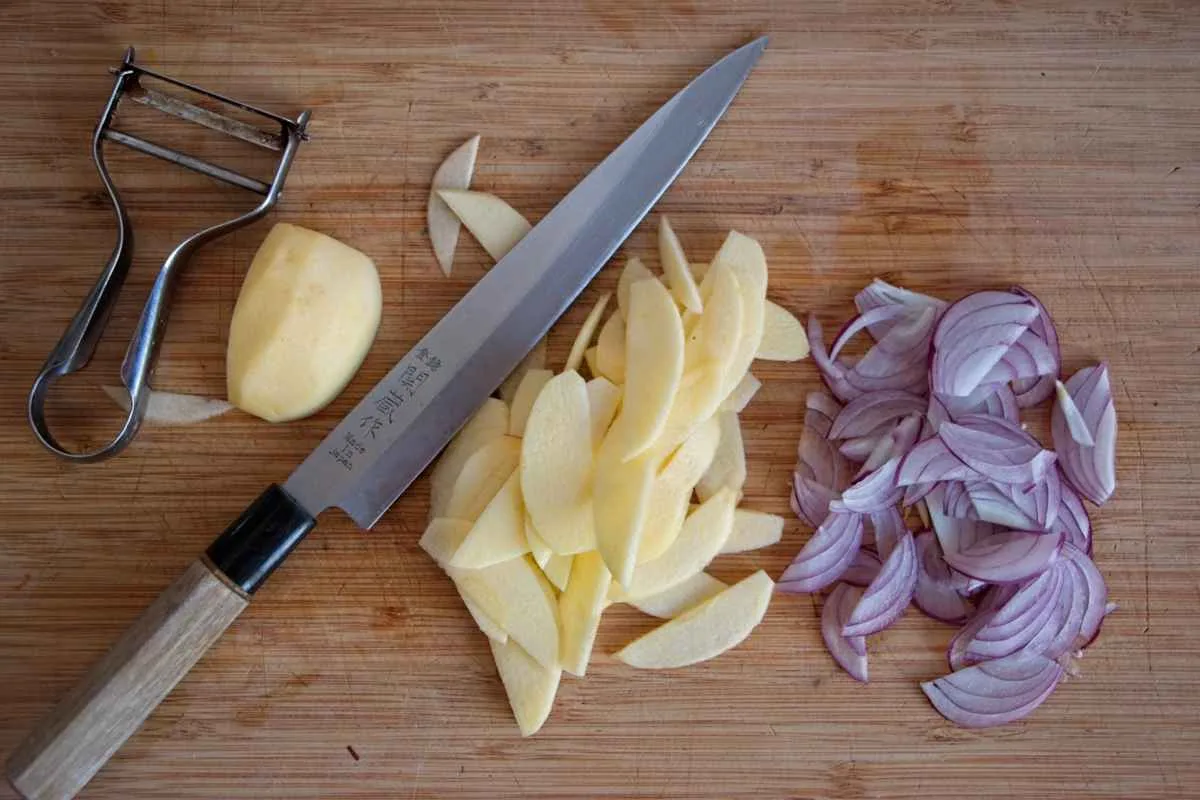 Sliced apples and red onion on a wooden board with a chef’s knife and peeler.
