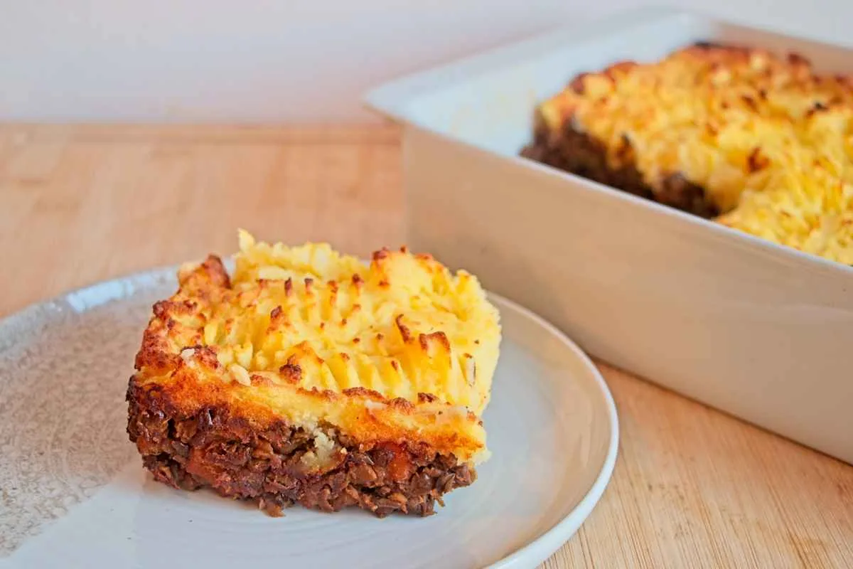 A slice of vegetarian cottage pie with porcini served on a plate, with the rest of the pie in the baking dish behind it