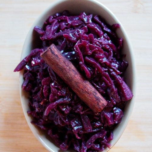 Bowl of slow-cooked red cabbage topped with a cinnamon stick on a wooden board.