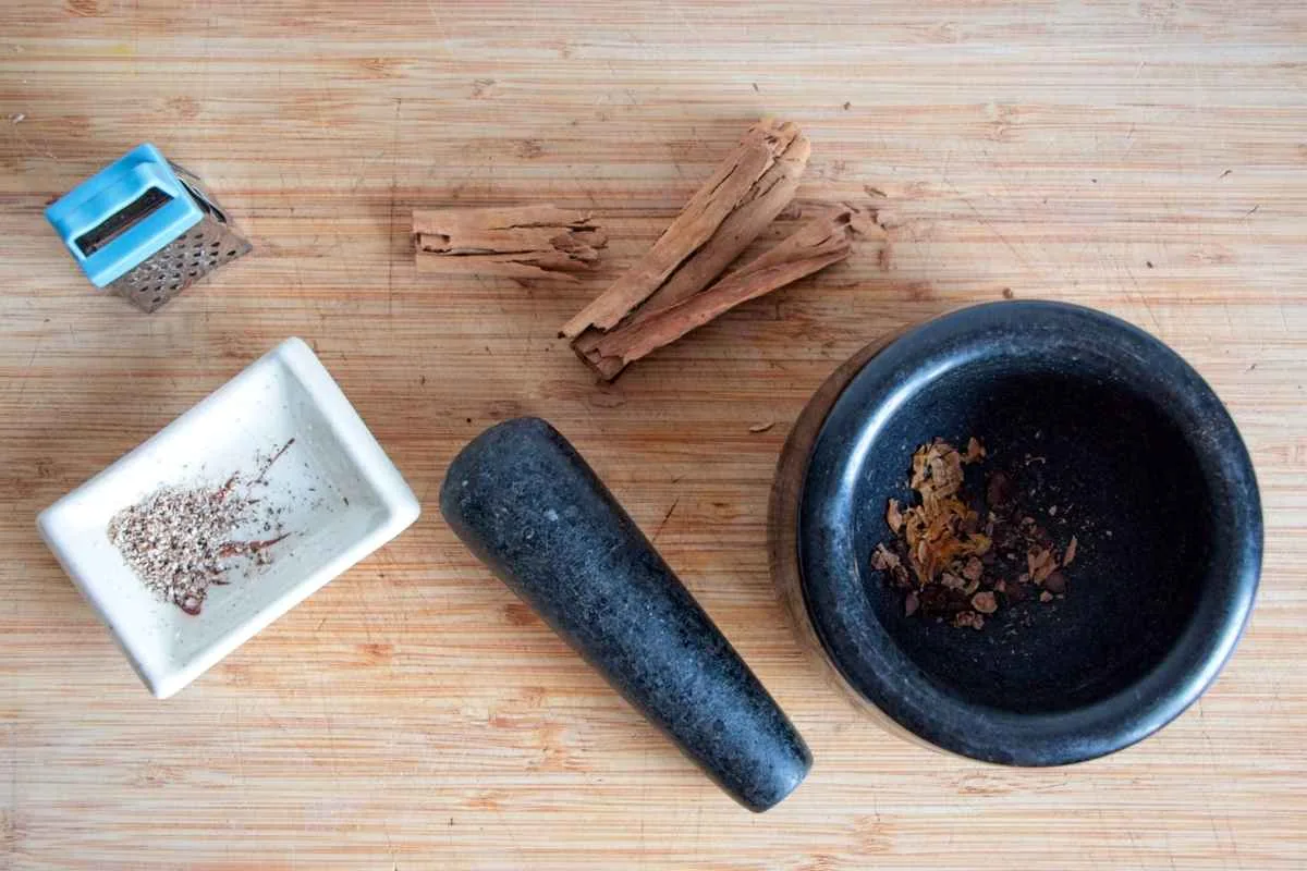 Toasted whole spices and nutmeg being crushed with a mortar and pestle on a wooden board.