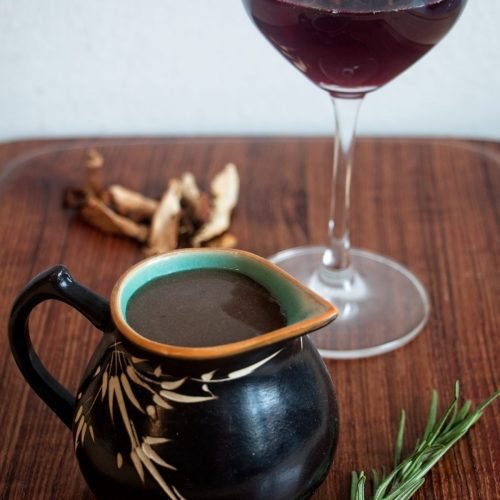 A jug of vegan red wine gravy on a wooden board with dried ceps, rosemary, and a glass of red wine in the background.