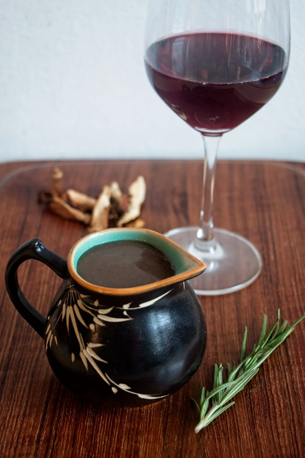 A jug of vegan red wine gravy on a wooden board with dried ceps, rosemary, and a glass of red wine in the background.
