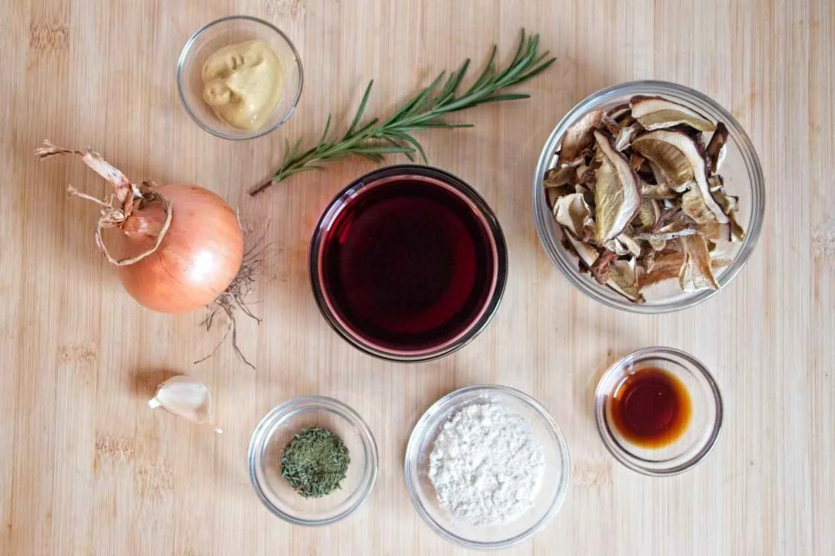 Ingredients laid out for vegan red wine gravy, including dried ceps, onion, garlic, rosemary, flour, mustard, soy sauce, and red wine.