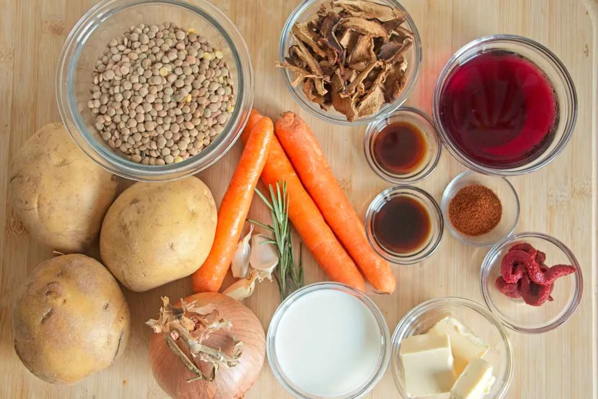 Ingredients for a vegetarian cottage pie with porcini laid out on a board, including lentils, carrots, potatoes, onions, dried mushrooms, stock, and seasonings.