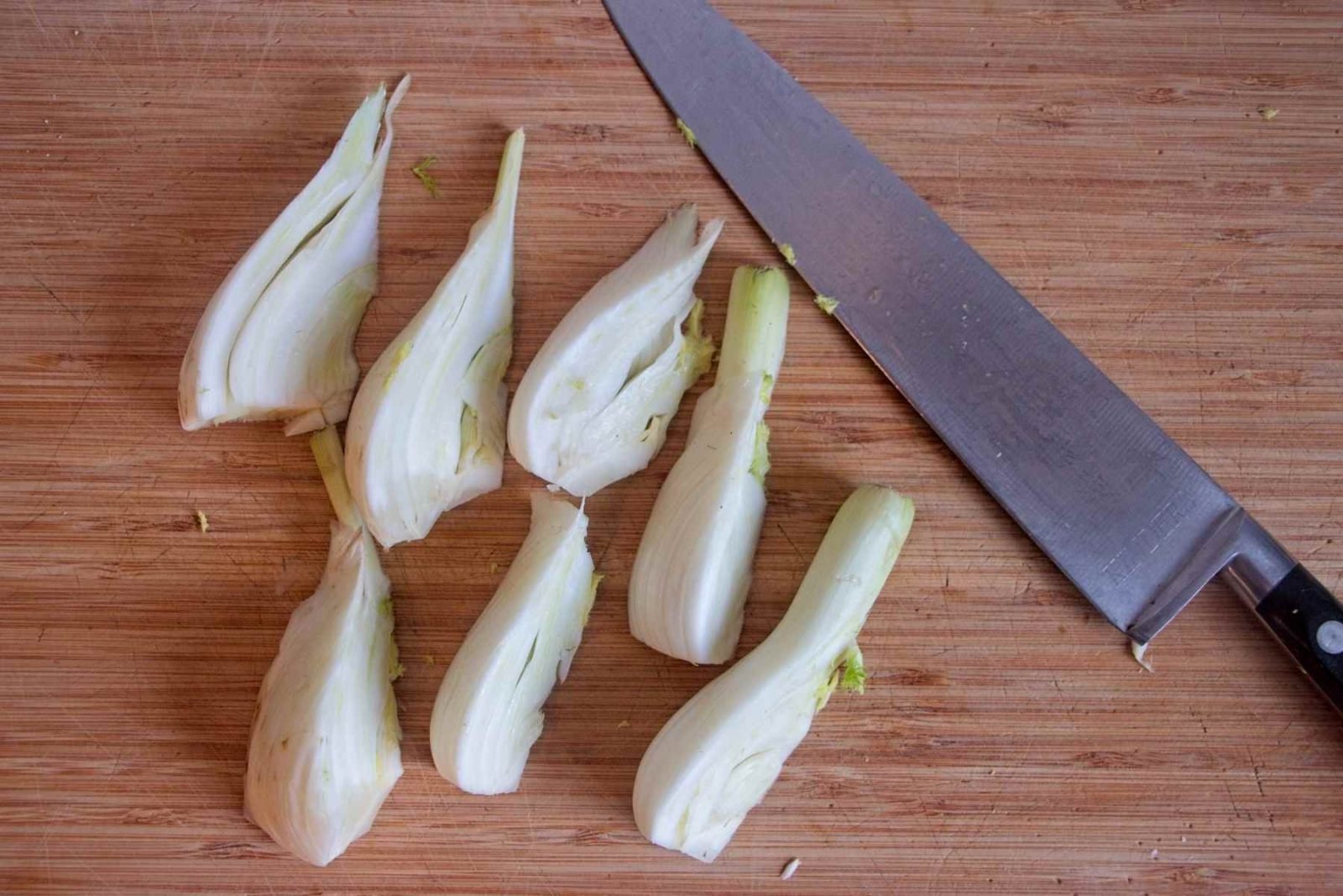 Fennel bulb cut into wedges on a wooden board with a knife, ready for cooking.
