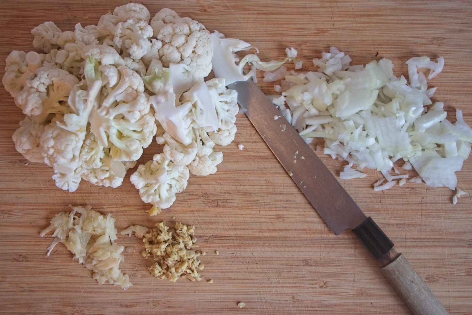 Cauliflower florets roughly chopped on a wooden board with a knife alongside.