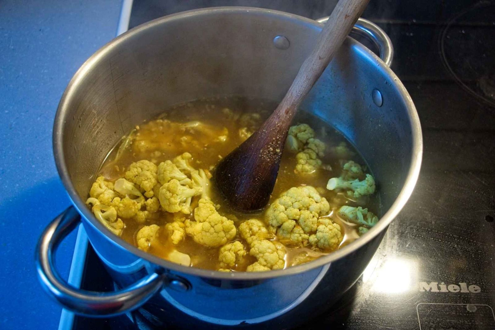 Cauliflower simmering in spiced stock in a large pot on the hob, with a wooden spoon stirring.