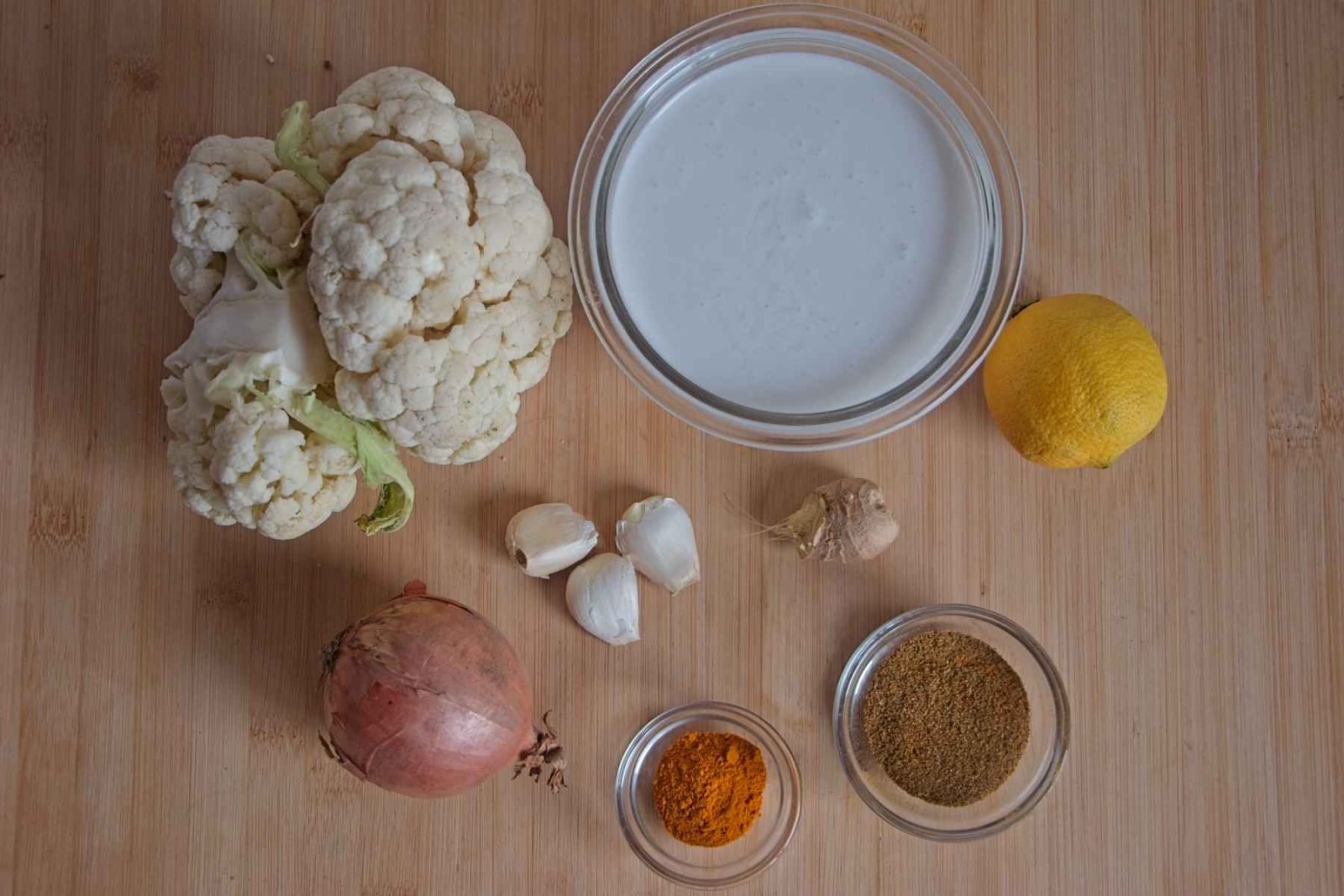 Ingredients for curried cauliflower soup arranged on a wooden board, including cauliflower, coconut milk, onion, garlic, spices, and lemon.