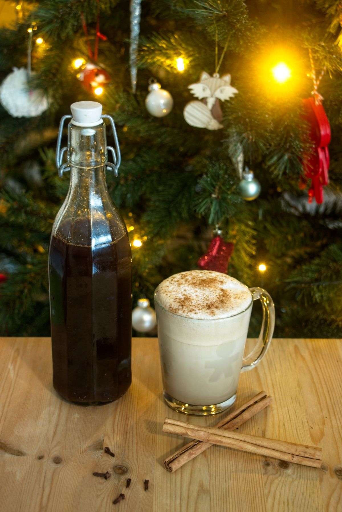 Gingerbread syrup in a bottle next to a spiced latte, photographed in front of a Christmas tree with warm festive lights.