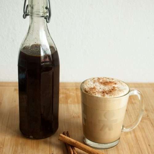 Gingerbread syrup in a glass bottle beside a spiced latte, with cinnamon sticks and cloves on a wooden board.