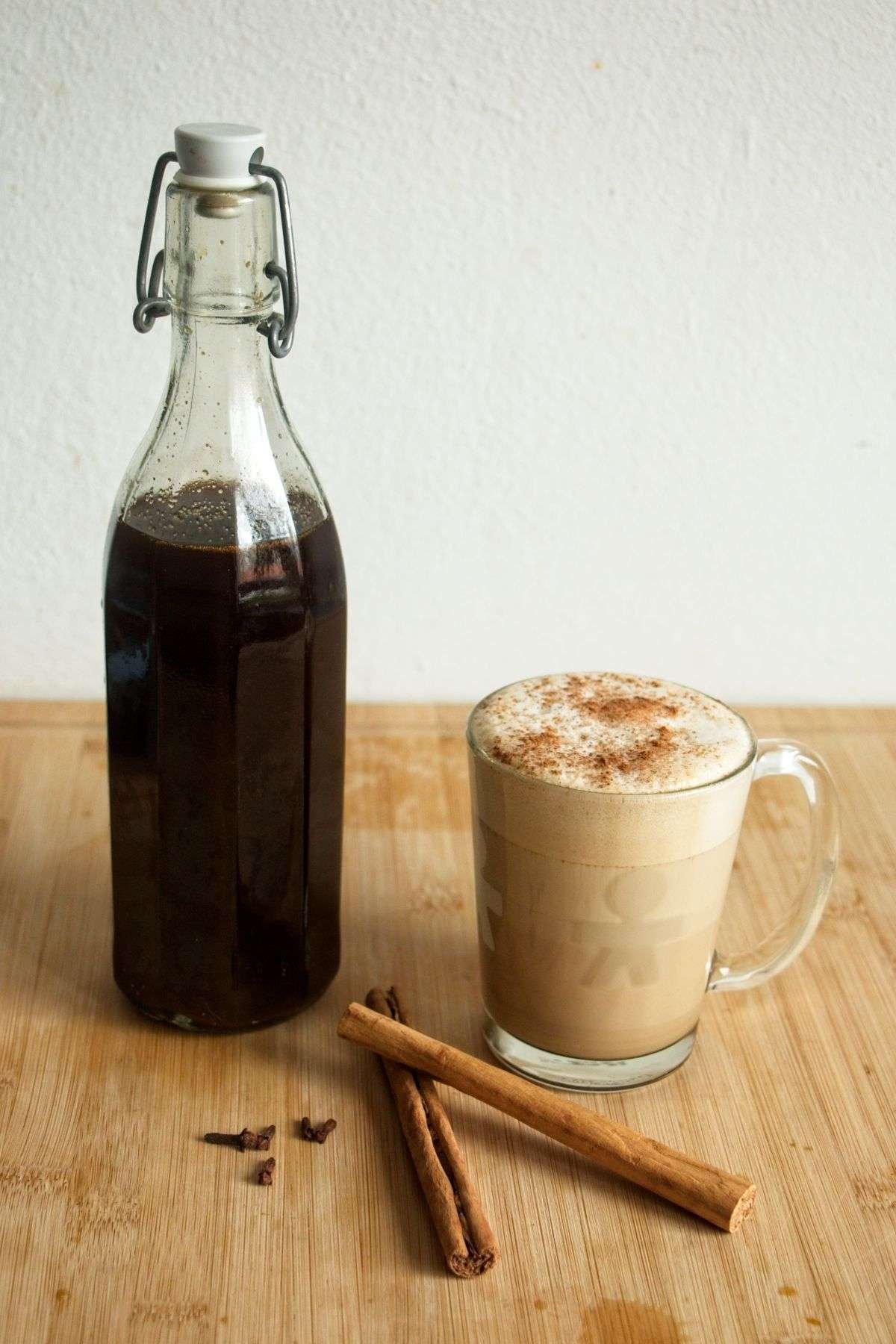 Gingerbread syrup in a glass bottle beside a spiced latte, with cinnamon sticks and cloves on a wooden board.