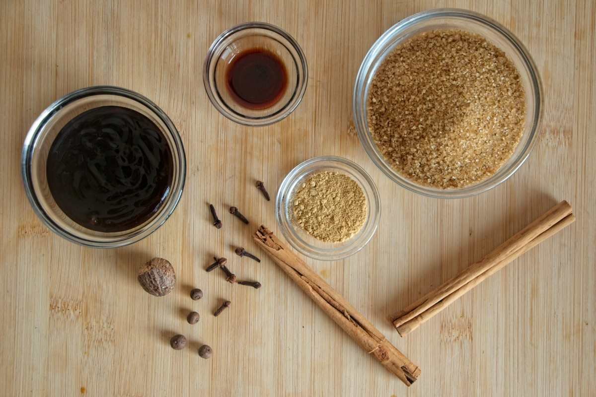 Gingerbread syrup ingredients laid out on a wooden board, including sugar, molasses, cinnamon sticks, cloves, allspice, and nutmeg.