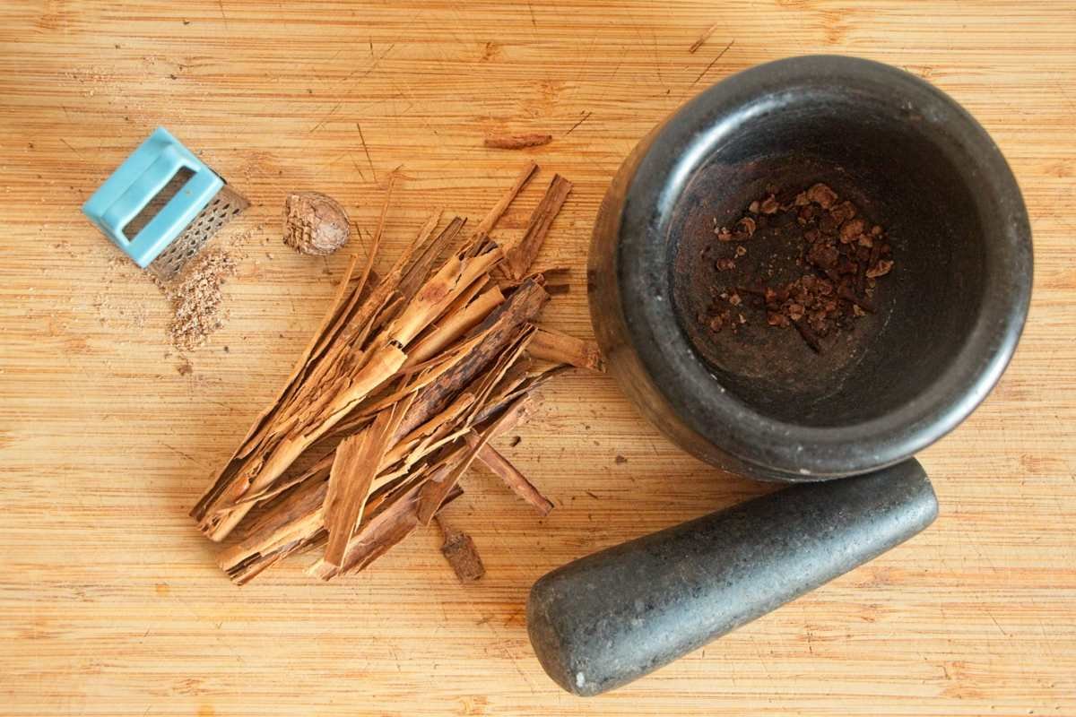 Crushed cloves in a mortar with cinnamon bark and freshly grated nutmeg on a wooden board.