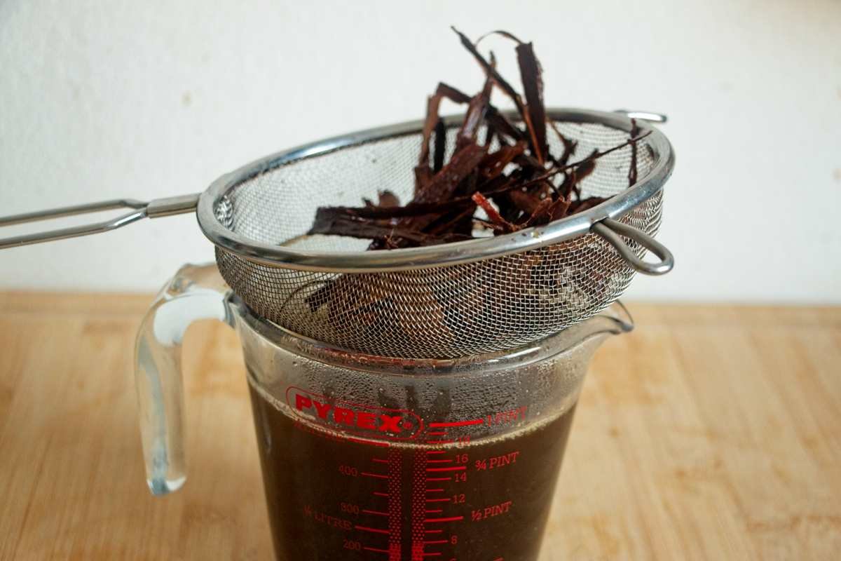 Warm spiced syrup being strained through a fine mesh sieve into a glass measuring jug.