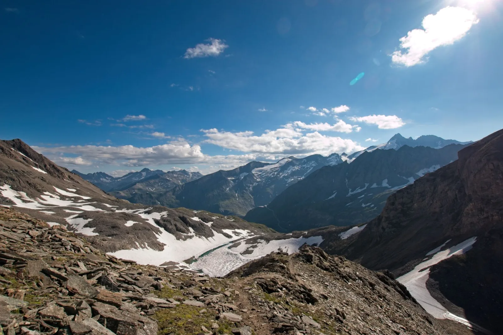 High-altitude landscape of the Glockner mountains featuring rugged rocky slopes, melting snowfields, and a wide blue sky with scattered clouds.