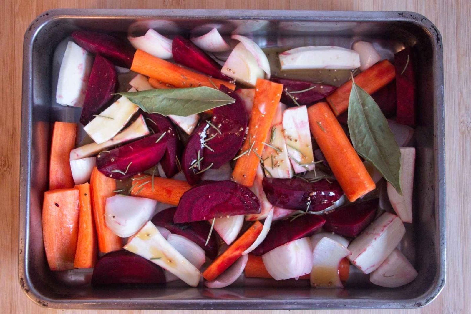 Roasting tray filled with carrots, parsnips, beetroot, and onion tossed with oil, mustard, bay leaves, and herbs before roasting