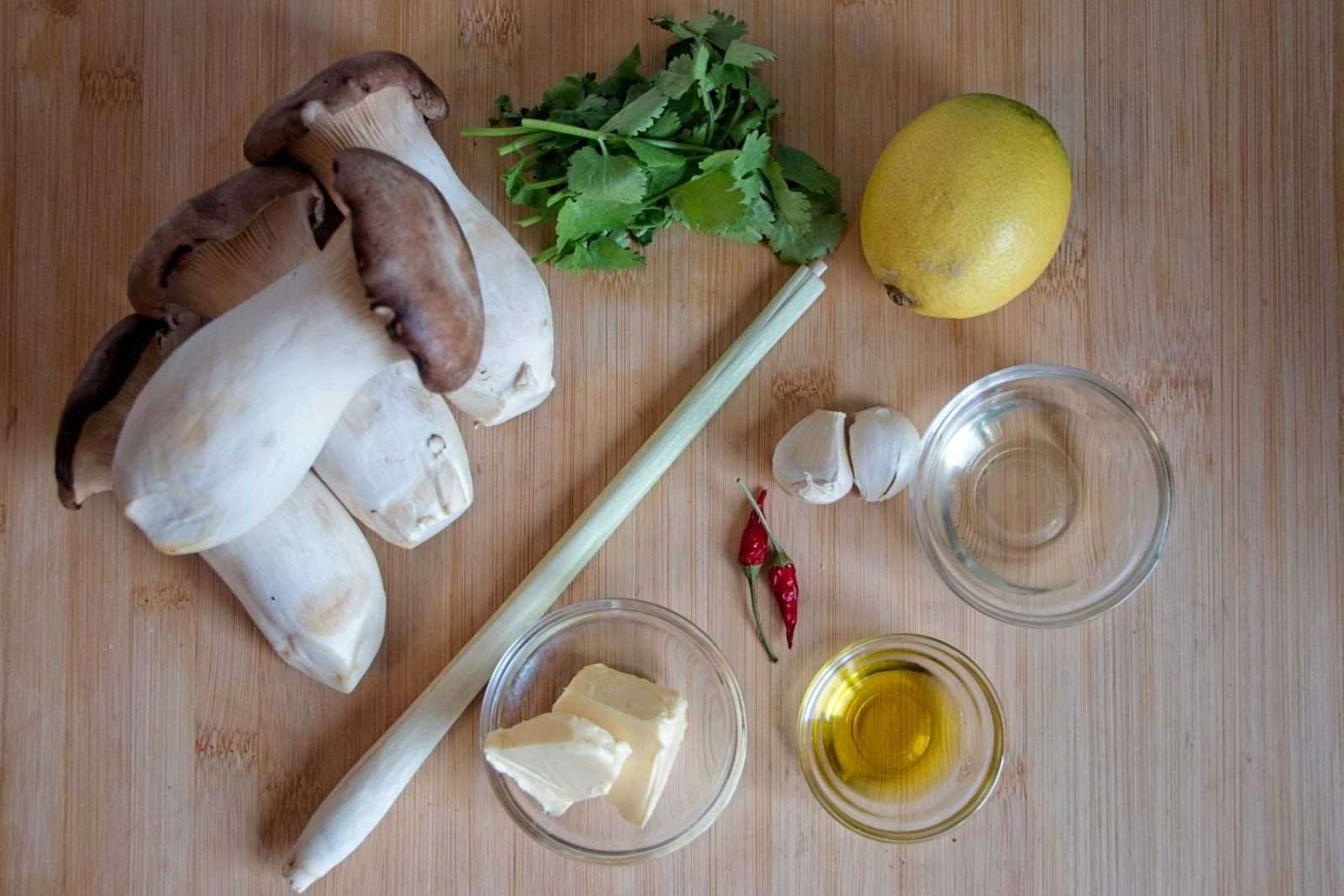 Ingredients for king oyster mushroom scallops, including mushrooms, lemongrass, lemon, garlic, chilli, butter, oil, and fresh herbs arranged on a wooden board.