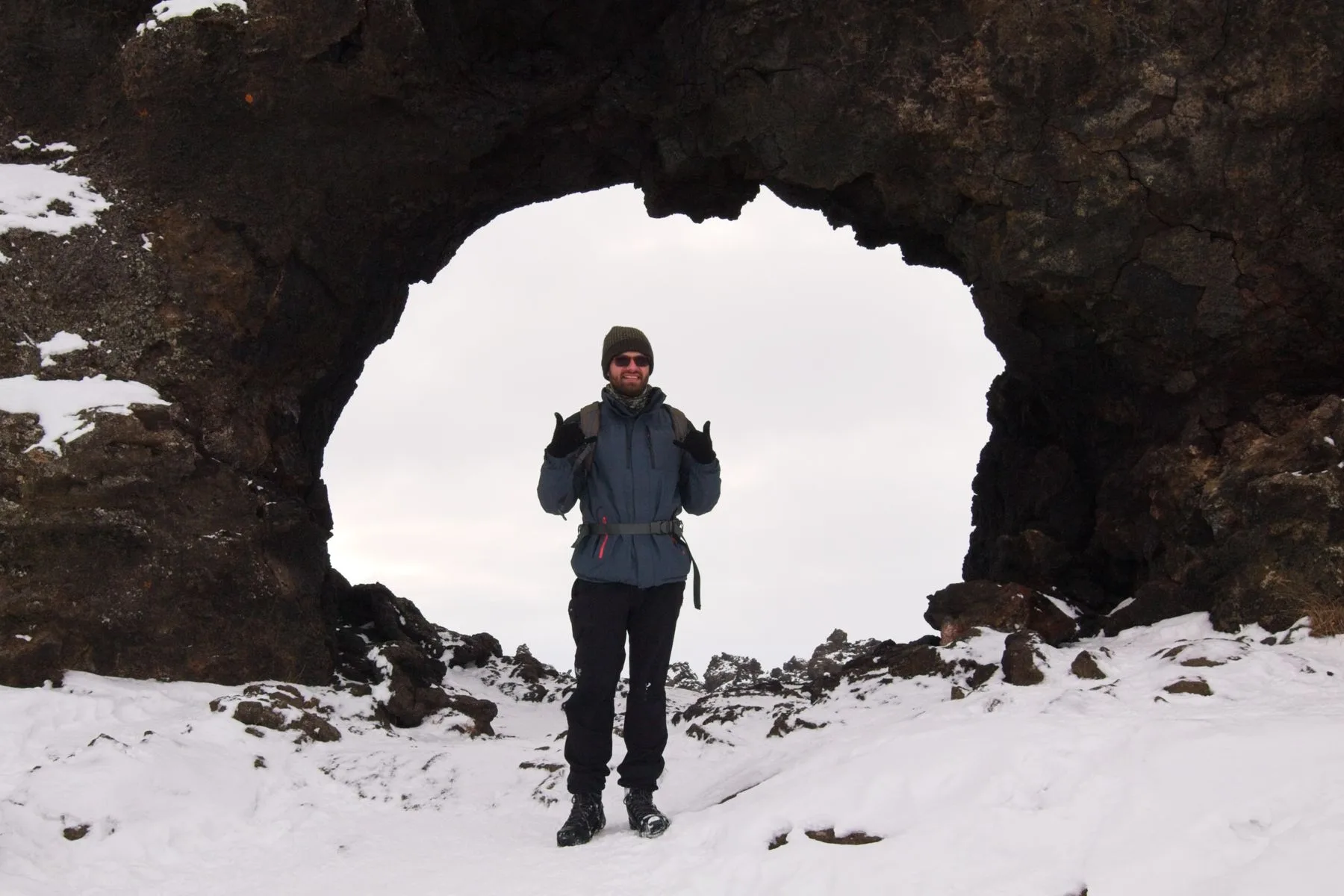 Hiker standing in front of a large natural rock arch in a snowy Icelandic landscape.