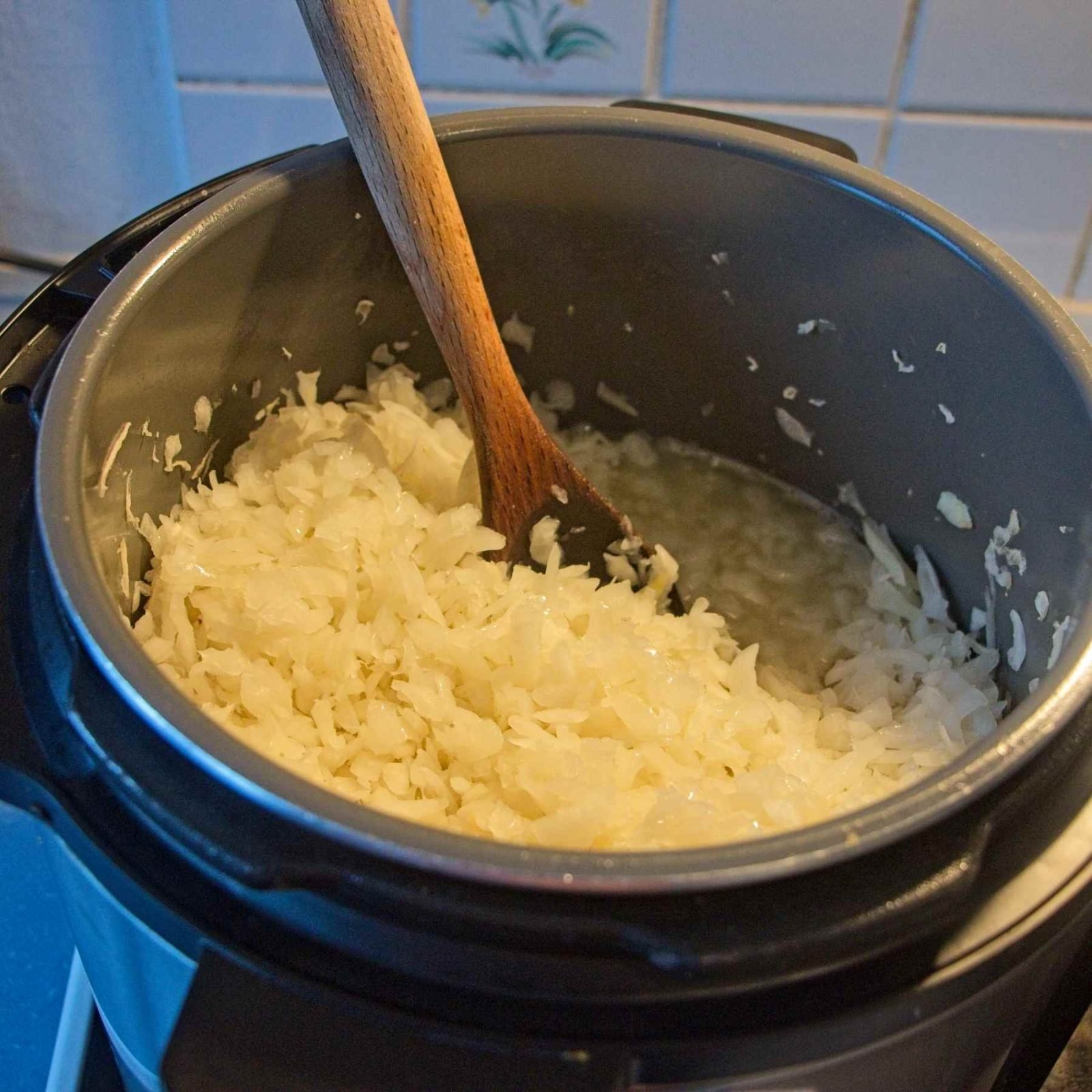 Chopped onions half way through cooking in an Instant Pot on slow cook mode, with a wooden spoon resting inside.