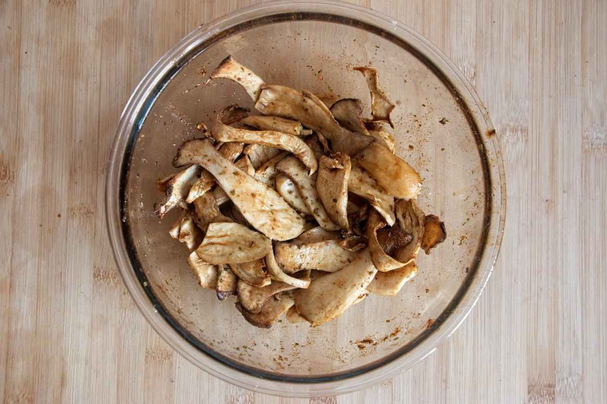 Sliced king oyster mushrooms coated in a dark savoury marinade resting in a glass bowl before draining and drying.