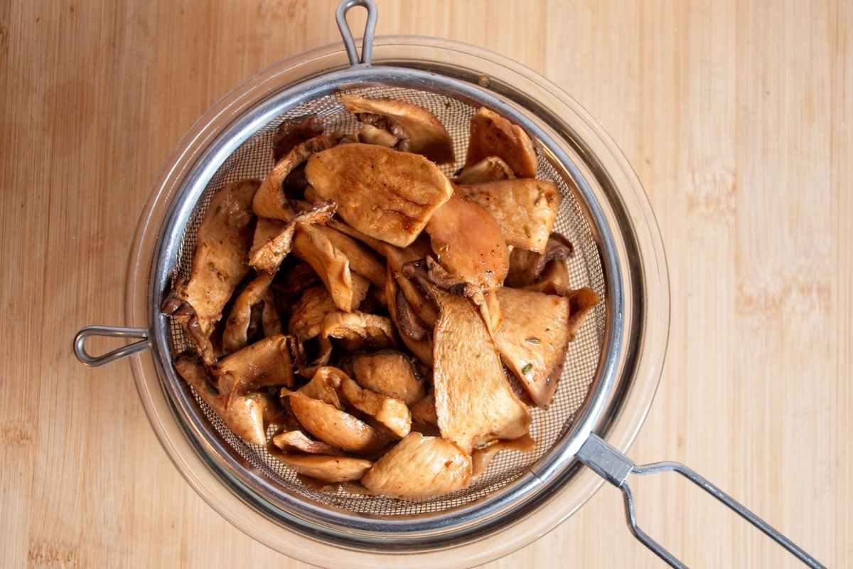 Marinated king oyster mushroom slices draining in a metal sieve over a bowl before being dried, coated evenly in a dark savoury marinade.
