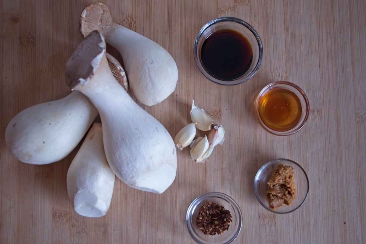 Ingredients for sticky king oyster mushrooms including whole king oyster mushrooms, garlic, soy sauce, miso paste, honey, and chilli flakes laid out on a board